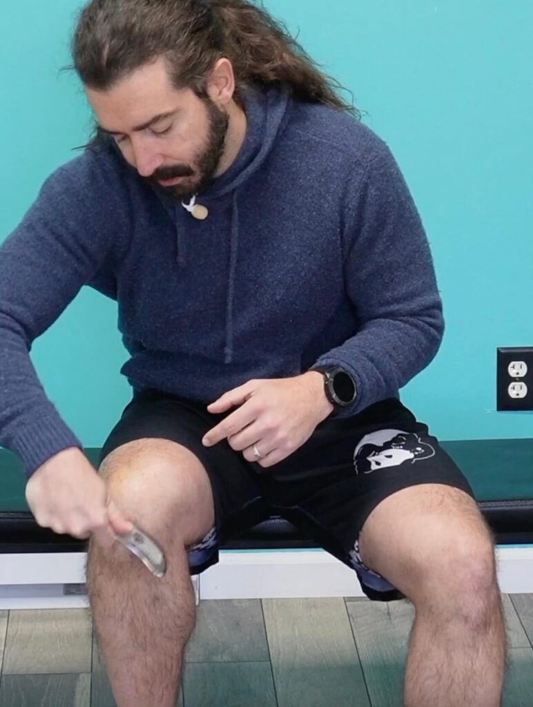 Dr. John seated on a bench using a steel tool to muscle scrape around his right knee - this is used for joint health to break up scar tissue